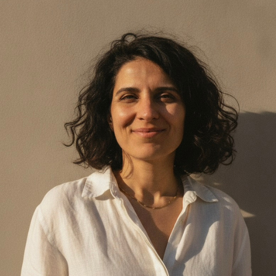 Ghazal’s headshot shows her smiling, with long dark curly hair, wearing a light gray blazer over a white top.